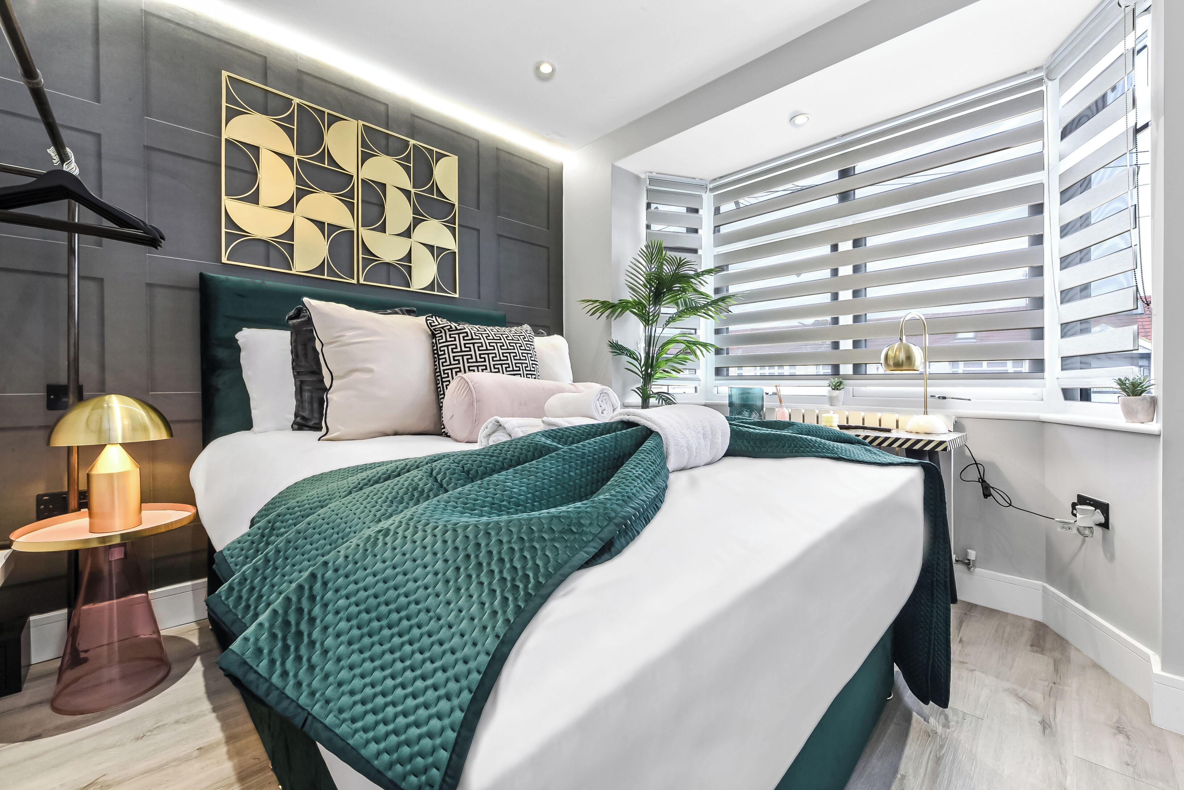 A modern bedroom with a large bed, a bedside table with a lamp, a decorative wall piece, and a window with blinds.