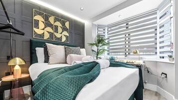 A modern bedroom with a large bed, a bedside table with a lamp, a decorative wall piece, and a window with blinds.