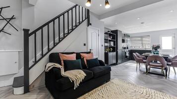 A modern living room with a black sofa, a staircase, and a dining area with pink chairs.