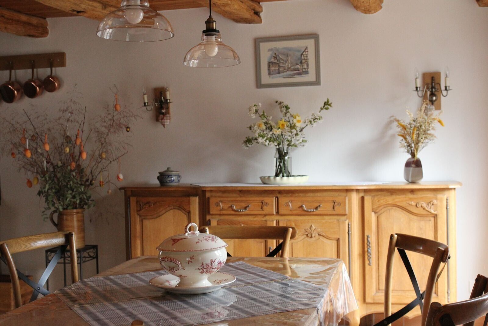 Zona pranzo con tavolo in legno, sedie, un vaso con fiori, un armadietto e utensili da cucina appesi.