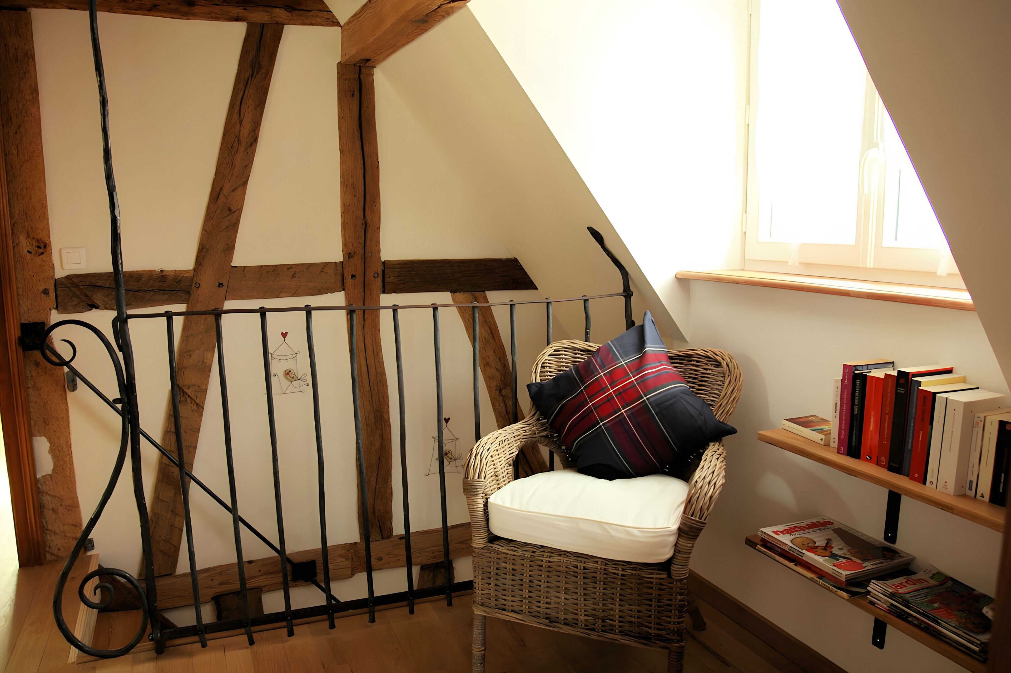 A room with a sloped ceiling, a wicker chair with a pillow, a metal railing, and a bookshelf with books.