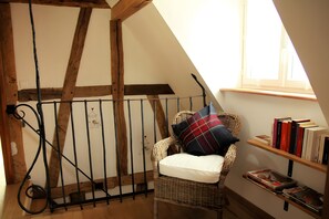 Una habitación con techo inclinado, una silla de mimbre con cojín, baranda de metal y estantería llena de libros.