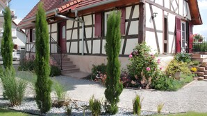 Una casa tradicional con entramado de madera y techo rojo, un camino de grava y un jardín con diversas plantas y flores.