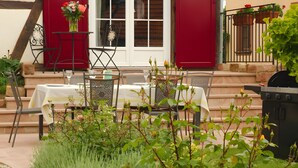 Un área de comedor al aire libre con una mesa lista para dos personas, rodeada de follaje y un frente de edificio rojo.
