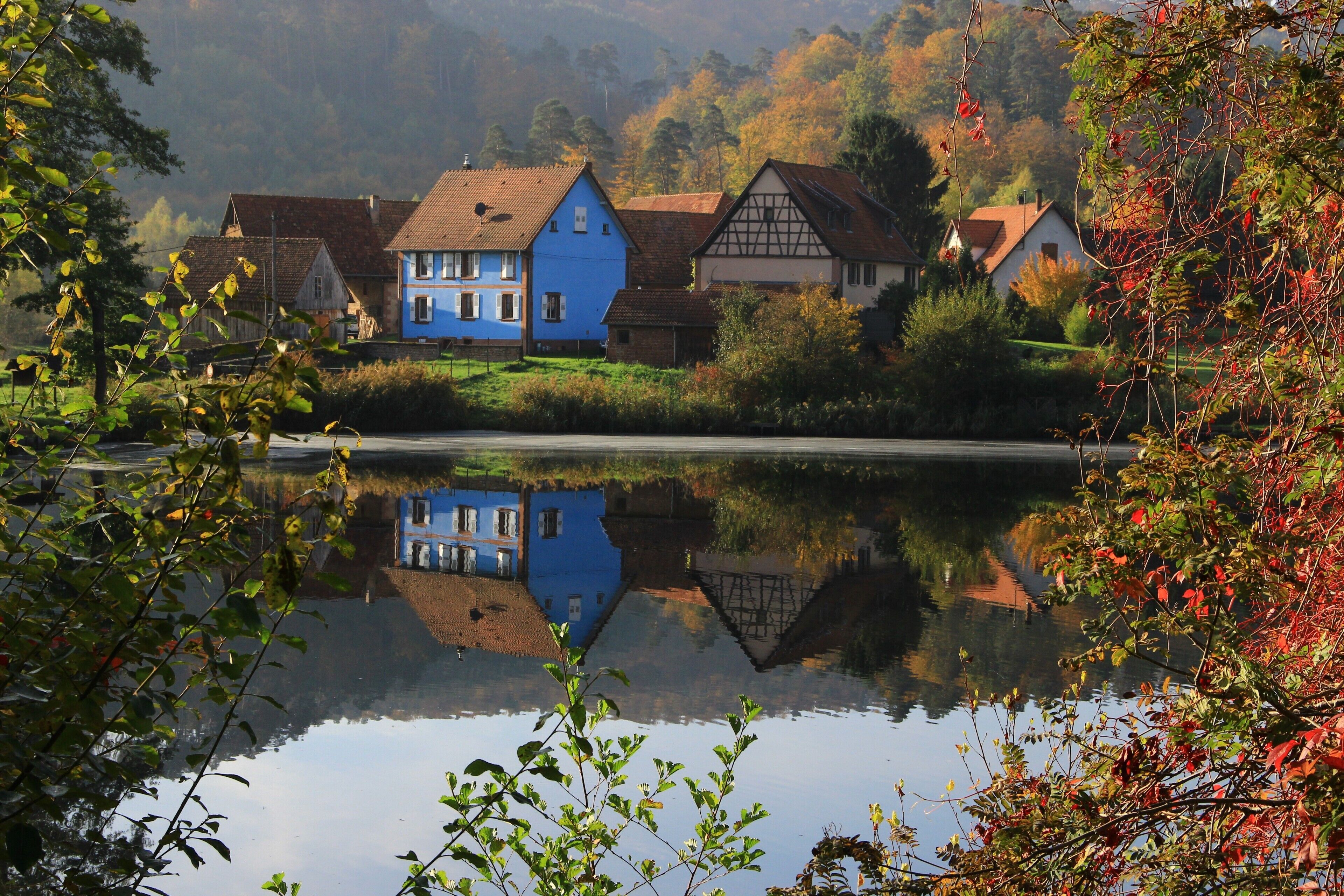 A tranquil lakeside village with traditional houses, a clear reflection in the water, and a forested backdrop.