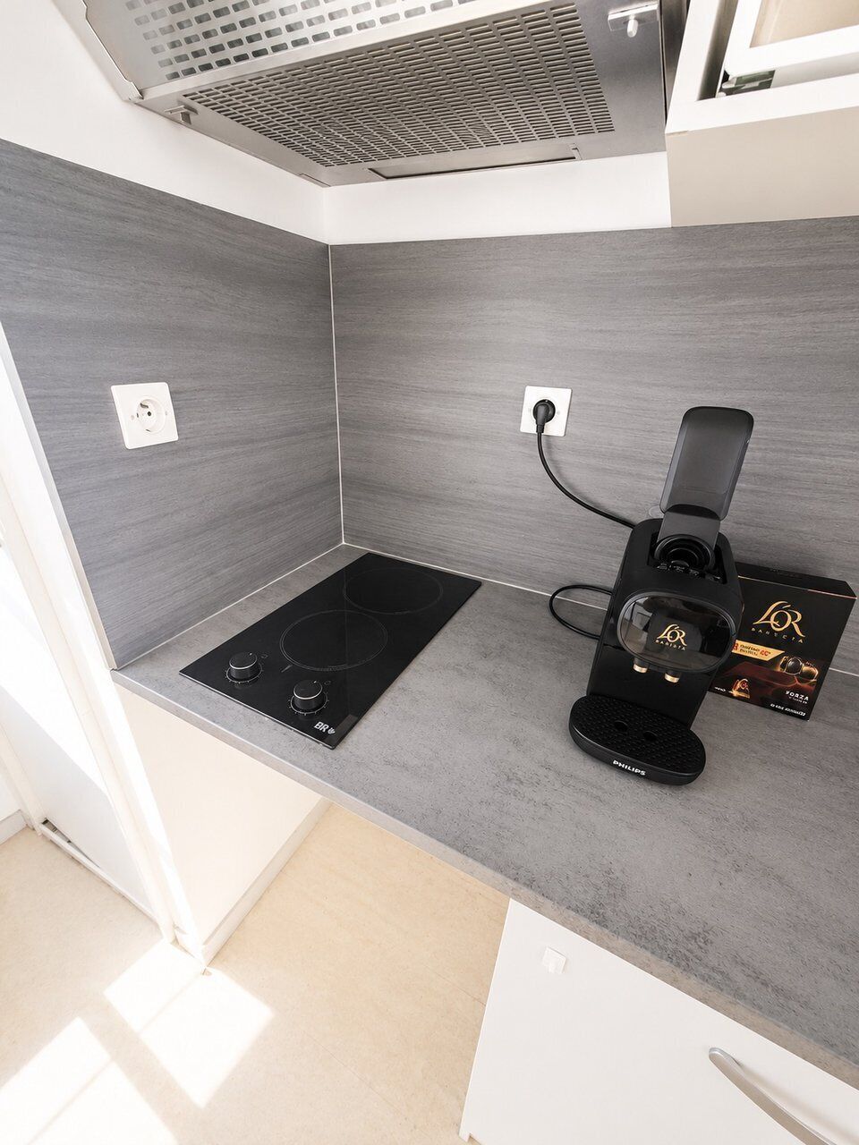 A compact kitchen with a black induction cooktop, a black coffee machine, and a white power outlet on the wall.