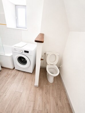 Ein kleines Badezimmer mit Waschmaschine, Toilette und einem kleinen Fenster.