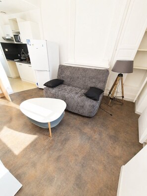 Ein kompakter Wohnbereich mit grauem Sofa, einem weißen Tisch, einer Lampe und einer Küchenzeile.