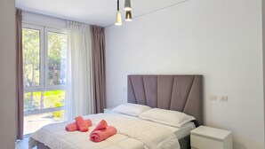 Un dormitorio con cama, almohadas, una mesita de noche y una ventana con cortinas.