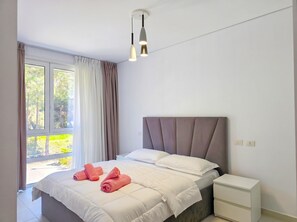 A bedroom with a bed, pillows, a bedside table, and a window with curtains. - Beachside Apartment 308 (Gjiri Lalzit)