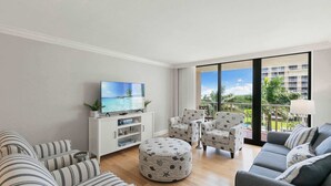 A living room with a TV, striped sofas, ottoman, and chairs, large windows with a view of buildings and greenery. - Remodeled Gulf-Front 2BR + Pool (Marco Island)