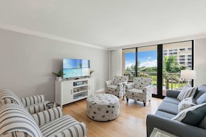 Ein Wohnzimmer mit Fernseher, gestreiften Sofas, Pouf und Sesseln, große Fenster mit Blick auf Gebäude und Grünflächen.