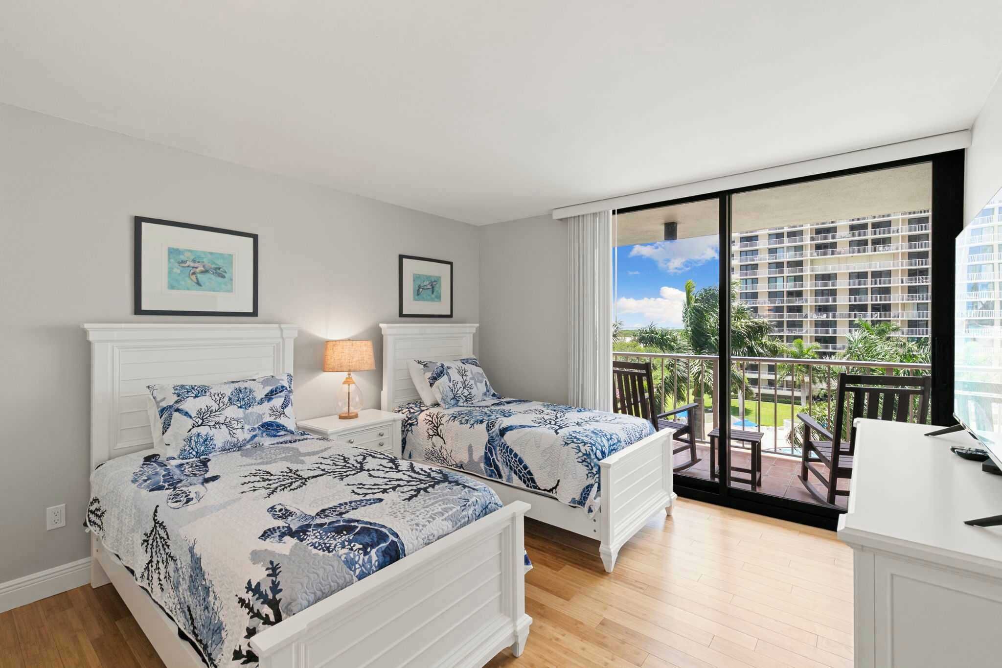 A bedroom with two beds, a desk, and a balcony with a view of a building and greenery.
