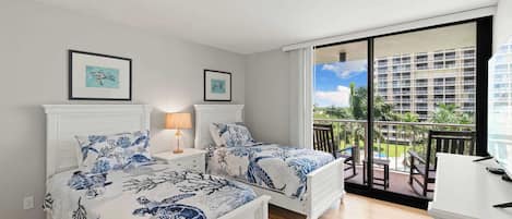 A bedroom with two beds, a desk, and a balcony with a view of a building and greenery.