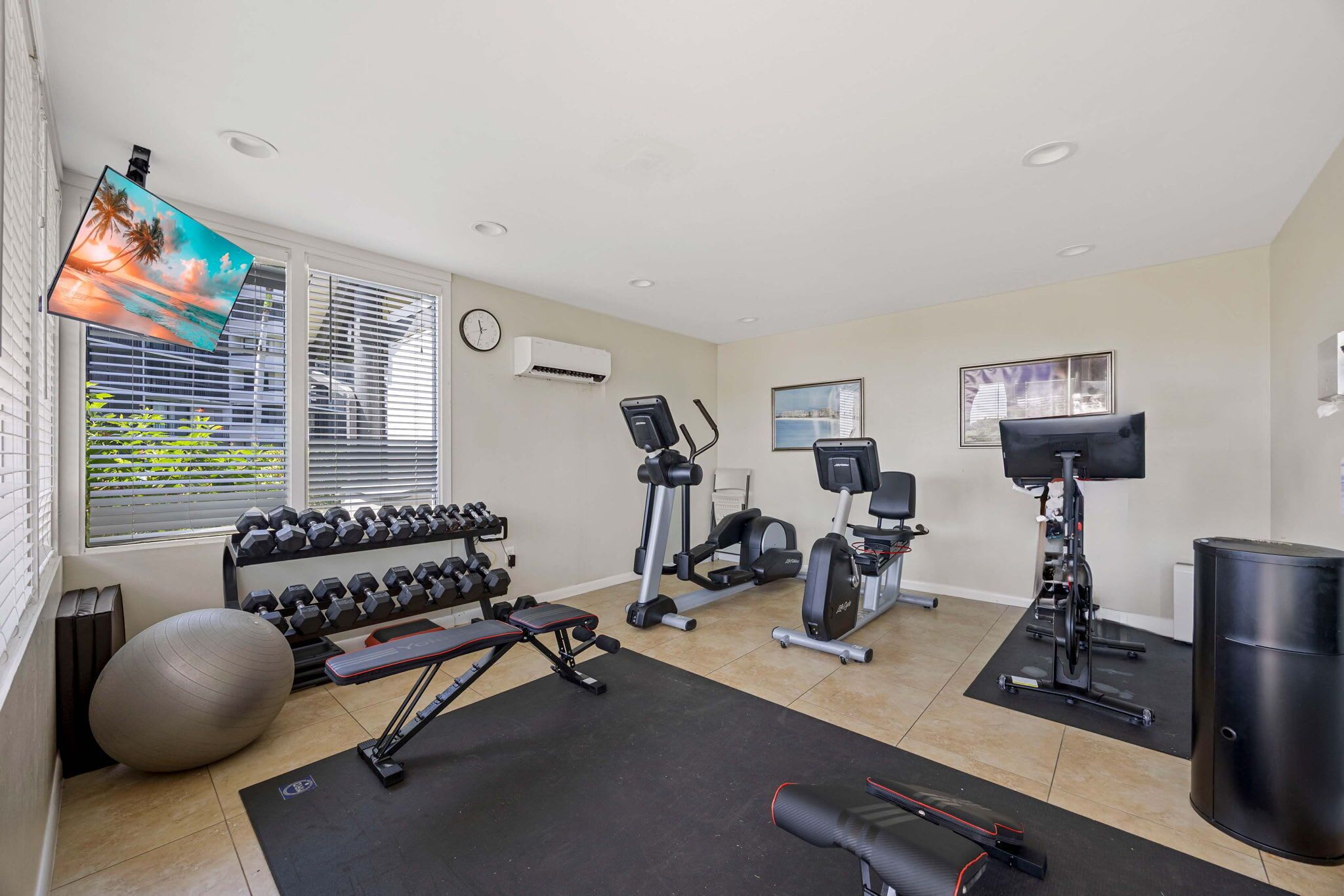 A home gym with exercise equipment, a TV, and a large window with blinds.