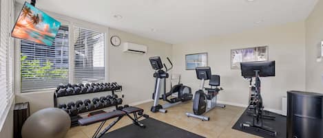 Un gimnasio en casa con equipo de ejercicio, un televisor y un ventanal con persianas.