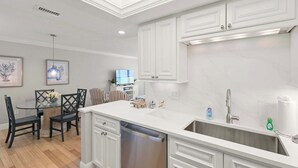 A modern kitchen with white cabinetry, a stainless steel sink, and a dining area with a glass table and chairs. - Remodeled Gulf-Front 2BR + Pool (Marco Island)
