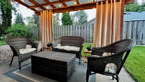 Eine Terrasse im Freien mit Korbmöbeln, einer Pergola mit Lichterketten und einem Holzzaun im Hintergrund.