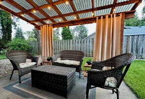 Un patio al aire libre con mobiliario de mimbre, una pérgola con luces colgantes y una valla de madera al fondo.