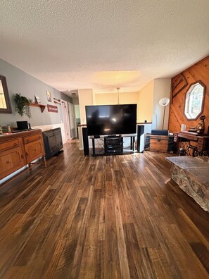 A living room with a flat-screen TV, wooden flooring, a stone accent piece, and a wooden wall with a window. - Beautiful Lake House Minutes from Lake Oroville (Oroville)