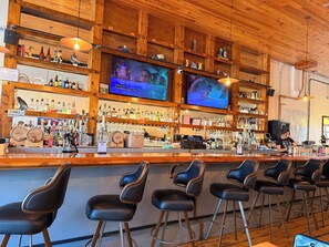 Un bar con estantes de madera llenos de botellas y vasos, dos televisores montados en la pared y varios taburetes de bar.