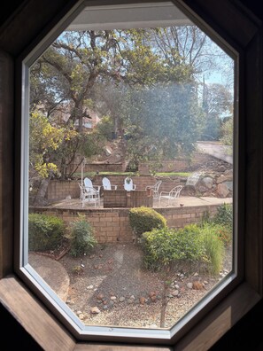 Vista a través de una ventana octogonal de un patio con sillas blancas, un muro de ladrillo y una variedad de plantas.