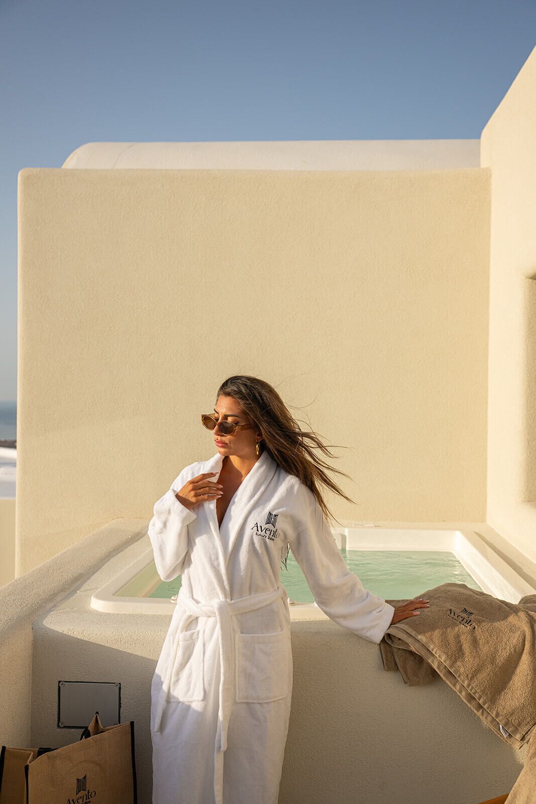 A person in a white robe standing on a balcony with a pool, a clear sky, and a beach in the background.