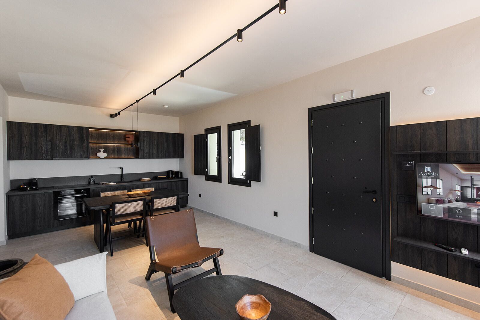 A modern kitchen with dark cabinetry, a dining area with a leather chair, and a flat-screen TV mounted on the wall.