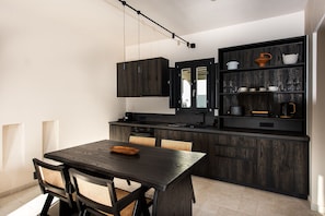 A modern kitchen with dark wood cabinetry, a built-in oven, and a dining area with a dark wooden table and chairs. - Avento Luxury Suites (Santorini)