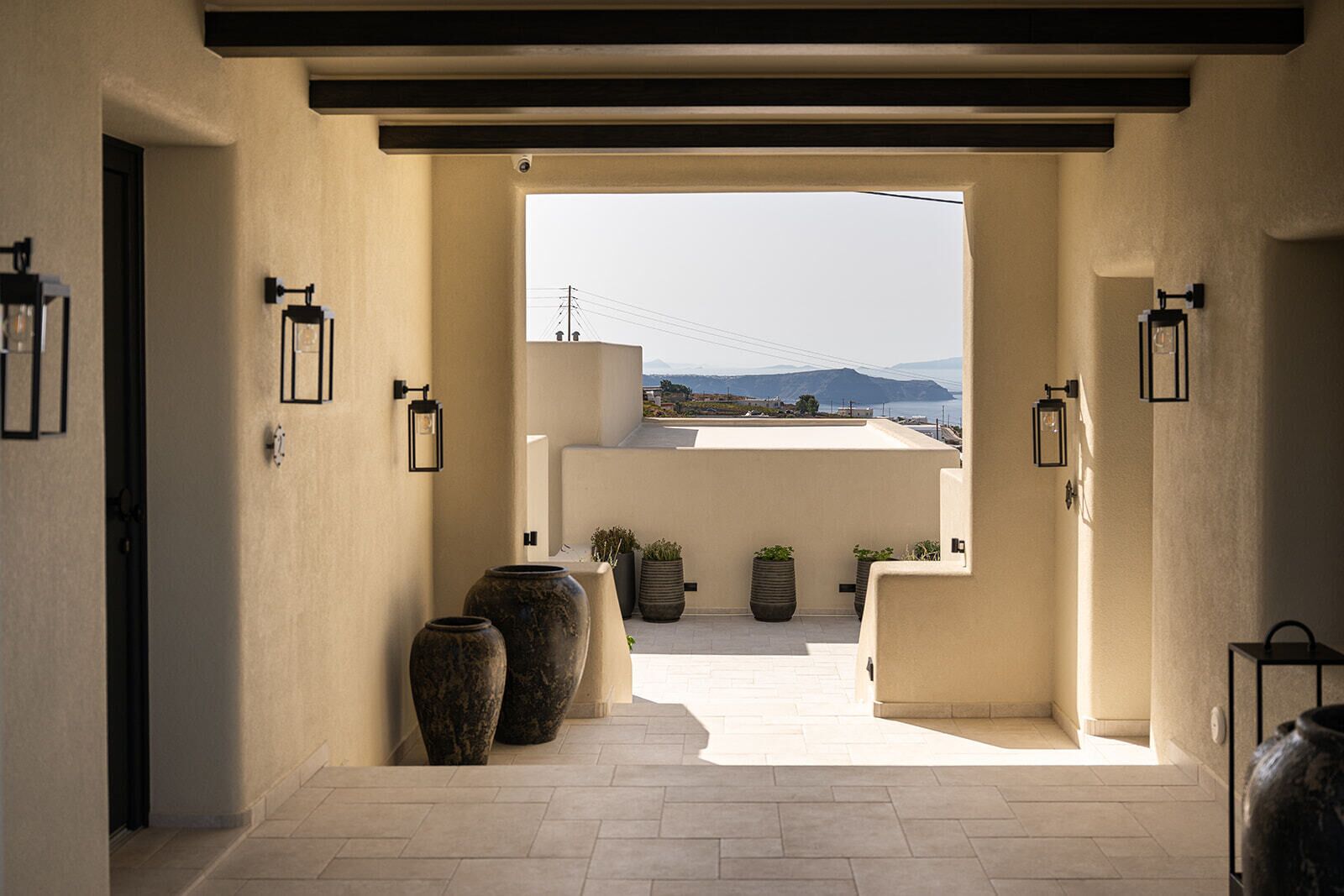 A corridor with a view of the sky and distant landscape, leading to a rooftop terrace with potted plants and a clear horizon.