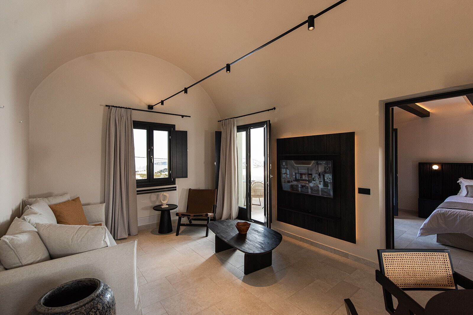 A modern hotel room with a curved ceiling, a sofa, a small table, a chair, a TV mounted on the wall, and a large window with curtains.