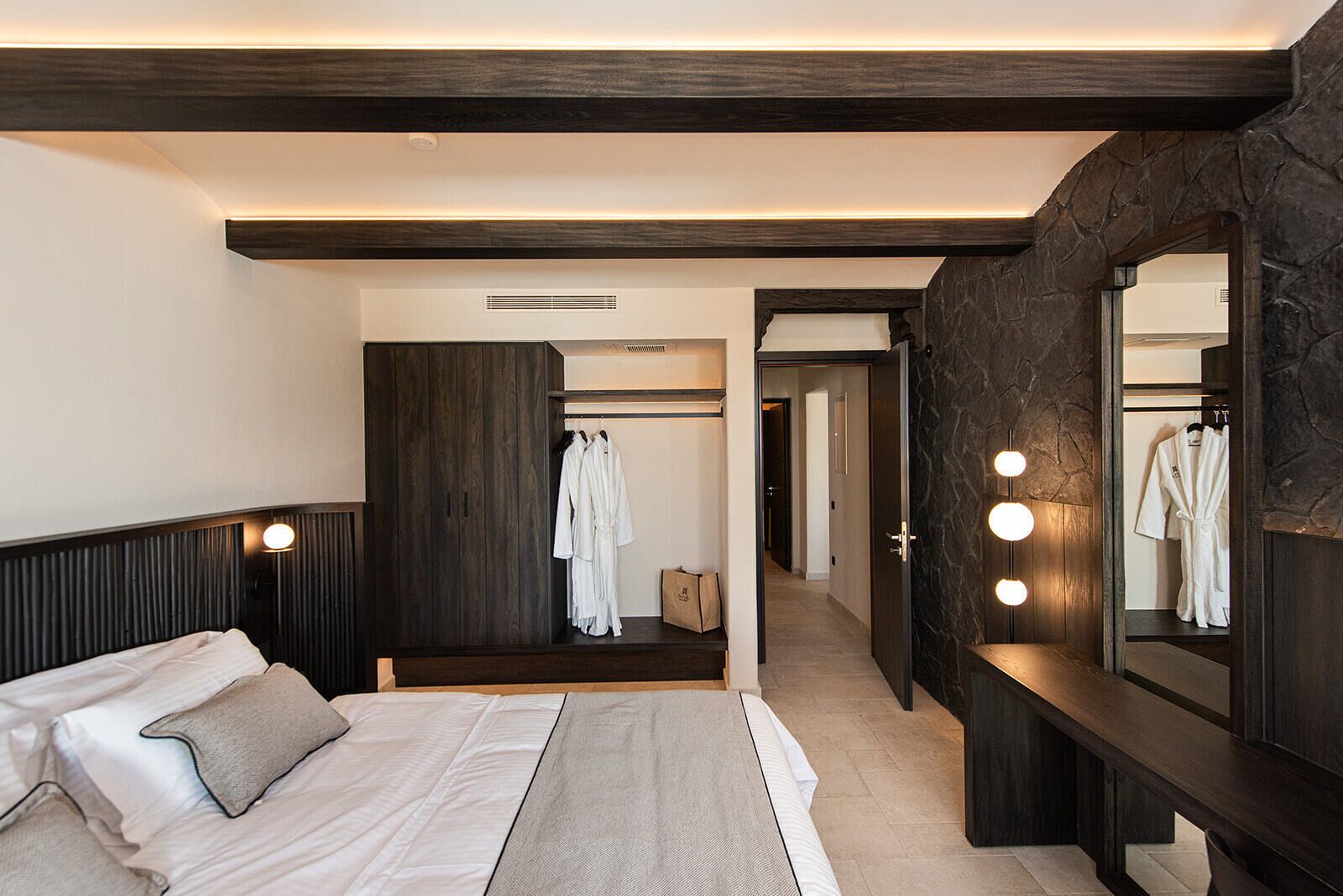 A bedroom with a large bed, a wardrobe, a mirror with lights, and a hallway leading to a bathroom.