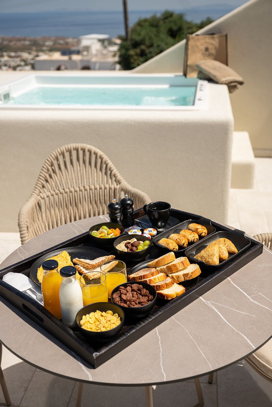 A breakfast tray with various foods and drinks on a table, with a pool and a chair in the background.