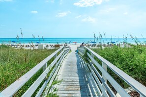 Ein hölzerner Steg führt zu einem Strand mit Liegestühlen und Sonnenschirmen.