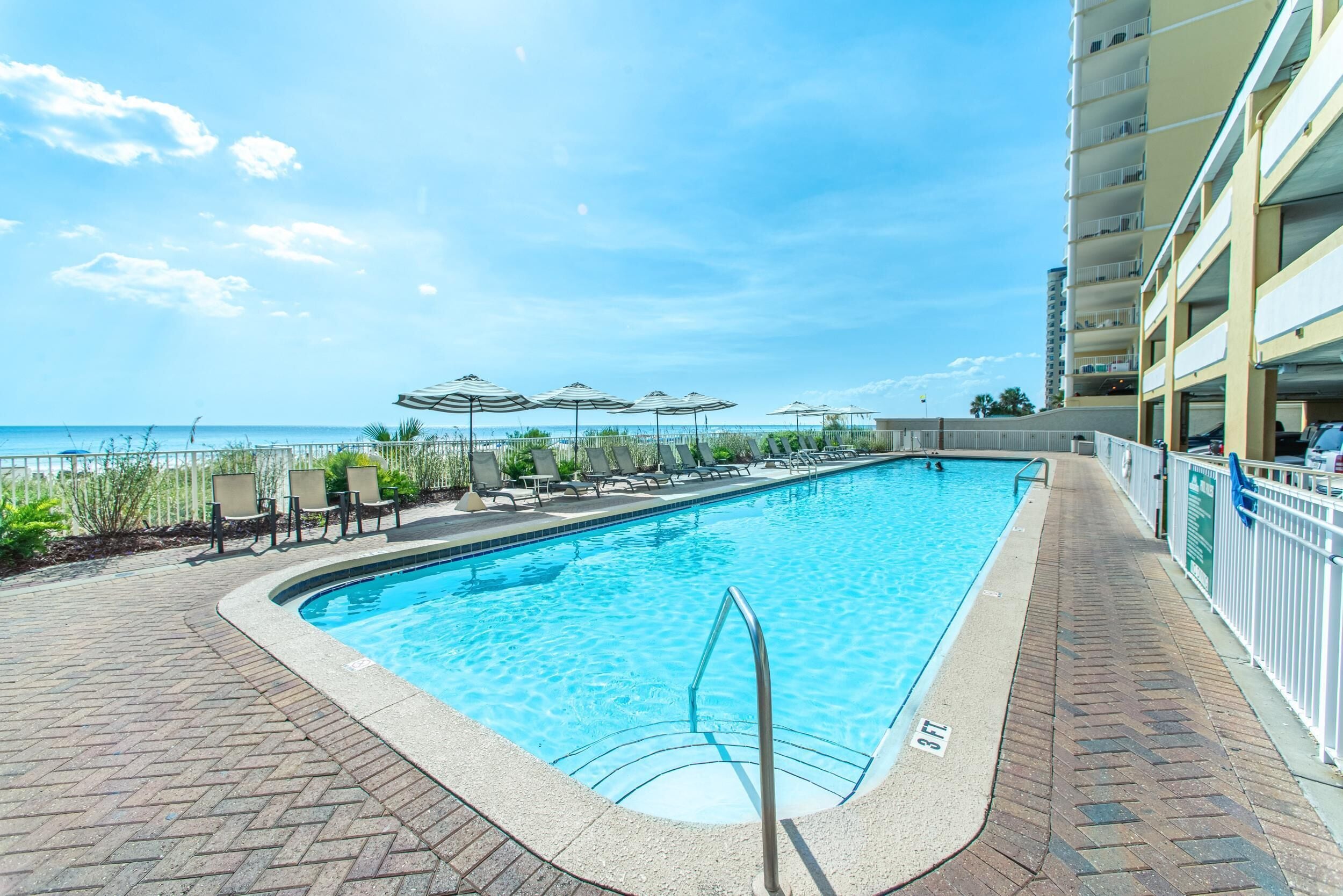 Ein rechteckiger Swimmingpool mit Metallleiter, umgeben von Liegestühlen und Sonnenschirmen, mit Blick auf das Meer und einem mehrstöckigen Gebäude im Hintergrund.