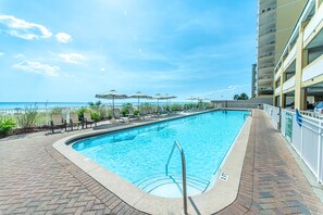 Ein rechteckiger Swimmingpool mit Metallleiter, umgeben von Liegestühlen und Sonnenschirmen, mit Blick auf das Meer und einem mehrstöckigen Gebäude im Hintergrund.