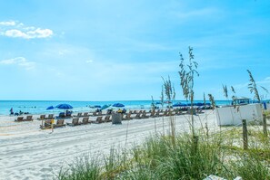 Ein Strand mit Reihen aus Holzliegen und blauen Sonnenschirmen, einem klaren Himmel und dem Meer im Hintergrund.