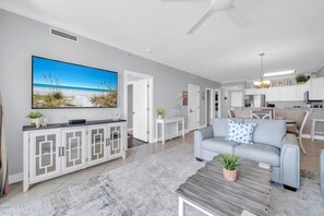 Een moderne woonkamer met een grote flatscreen-tv, een grijze bank, een salontafel en een keuken op de achtergrond.