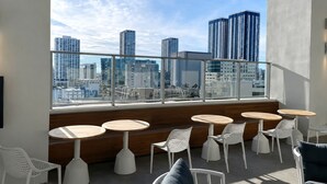 Una terraza en la azotea con mesas redondas y sillas blancas, con vistas a un paisaje urbano de edificios altos.