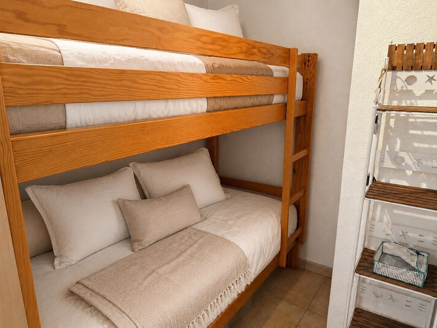 A bunk bed with a wooden frame, beige bedding, and a small shelf unit with decorative items.