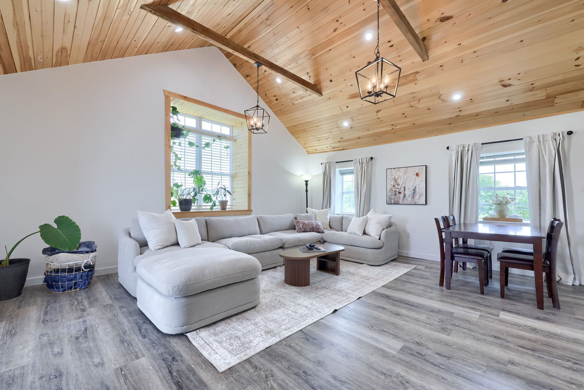 A modern living room with a sectional sofa, a dining table with chairs, wooden flooring, and a ceiling with exposed beams and pendant lighting.