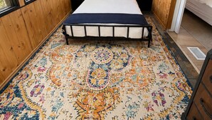 Makuuhuoneessa on puinen katto, kattovalaisin, kuviollinen matto sängyn alla ja yöpöytä, jossa on lamppu.