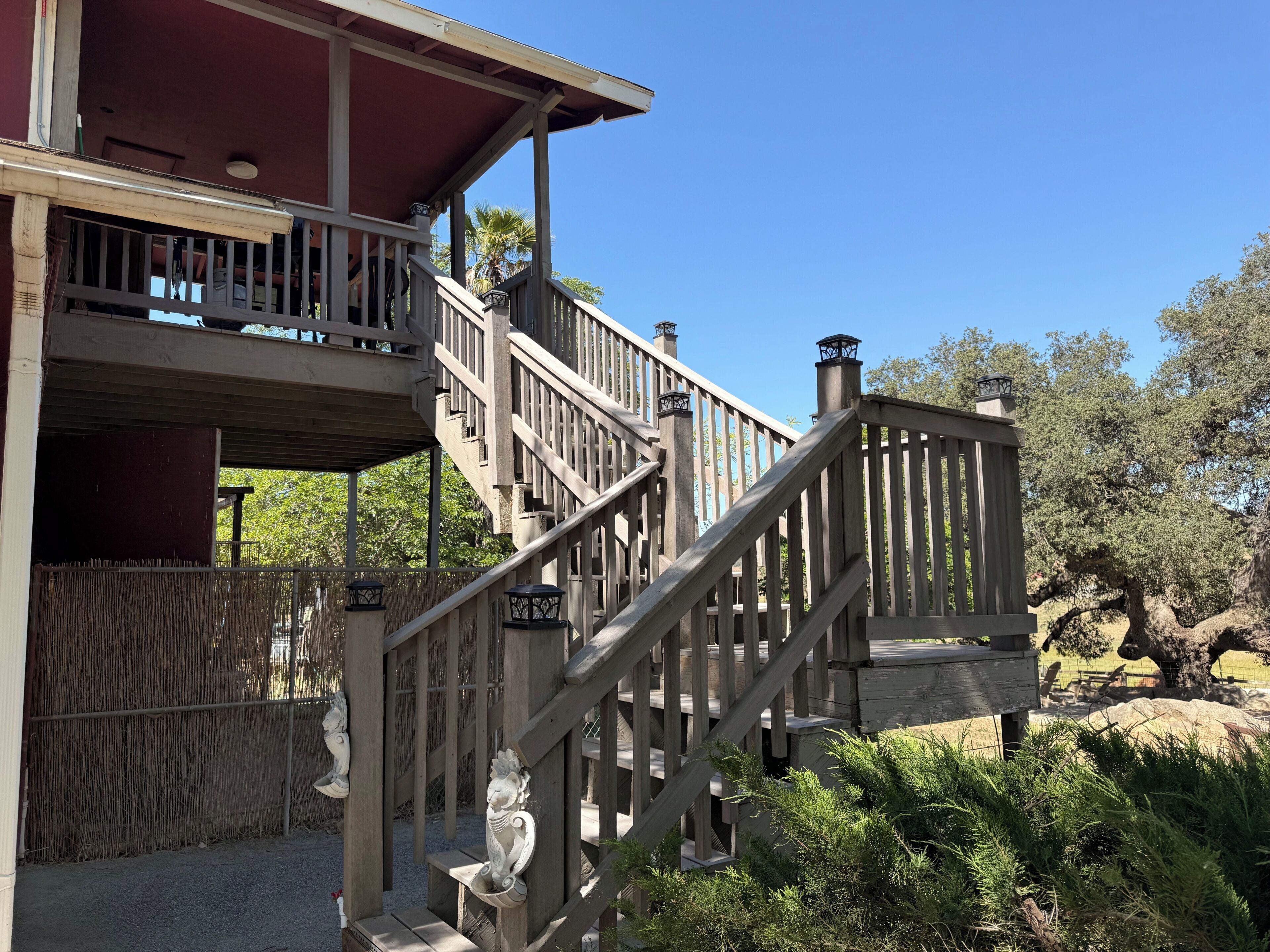 A wooden staircase leading to a covered upper level with a balcony, surrounded by greenery and trees.