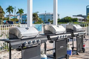 Drei mobile Grills auf Rädern, eine Gasflasche und ein Kühlkoffer auf einem befestigten Bereich mit Geländer und Palmen im Hintergrund.