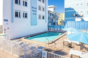 Ein Poolbereich mit Liegestühlen und Sonnenschirmen, ein Gebäude mit der Aufschrift 'BEL-AIR BEACH CLUB' und ein klarer, blauer Himmel.
