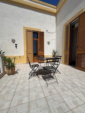 An outdoor patio with a table and chairs, a potted plant, and a door with wooden shutters. - Cozy oceanfront apartment with a garden and patio. (Santa Cesarea Terme)