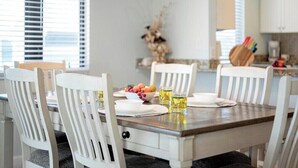 Un coin repas avec une table en bois dressée pour quatre personnes, des chaises blanches, un saladier rempli de fruits et deux verres contenant une boisson jaune.