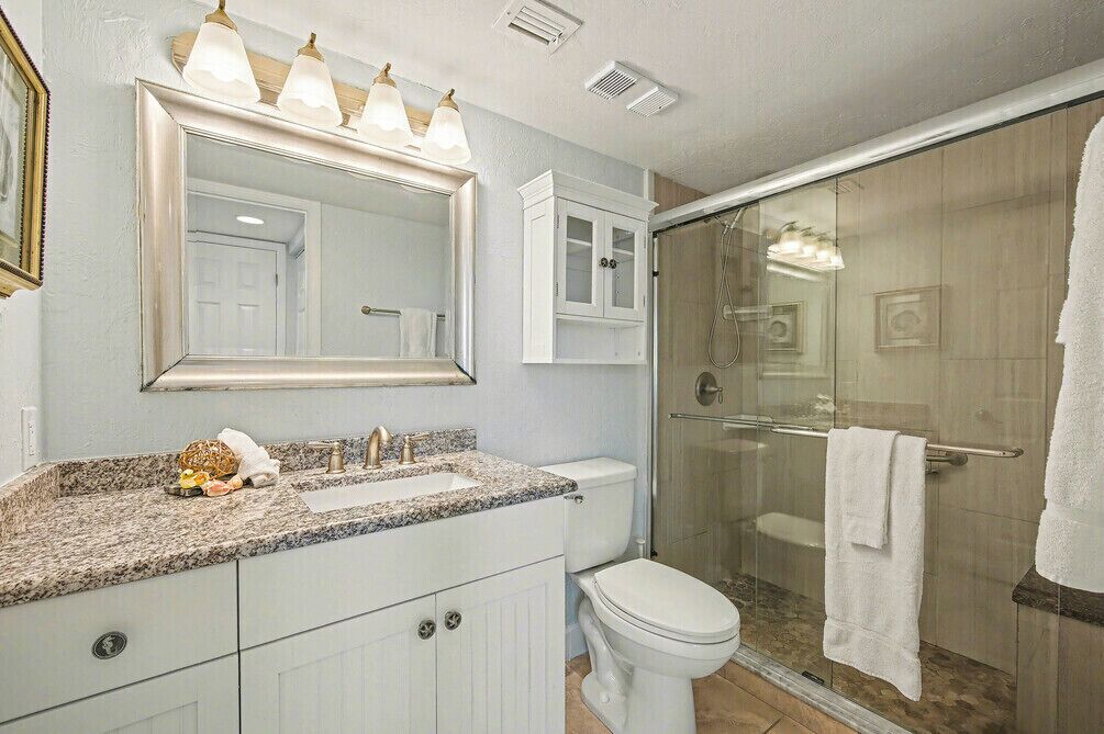 Une salle de bain dotée d’un comptoir en granit, de toilettes blanches, d’une douche en verre et d’un grand miroir avec un éclairage situé au-dessus.