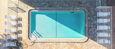 A rectangular swimming pool with a tiled deck surrounding it, equipped with a ladder and a diving board.