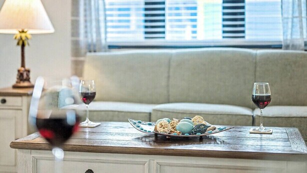 A dining table with two wine glasses and a plate of food, set against a sofa and a lamp in a room with a window. - Poolside 2BR Condo with Balcony near Beach Pier (Fort Myers Beach)